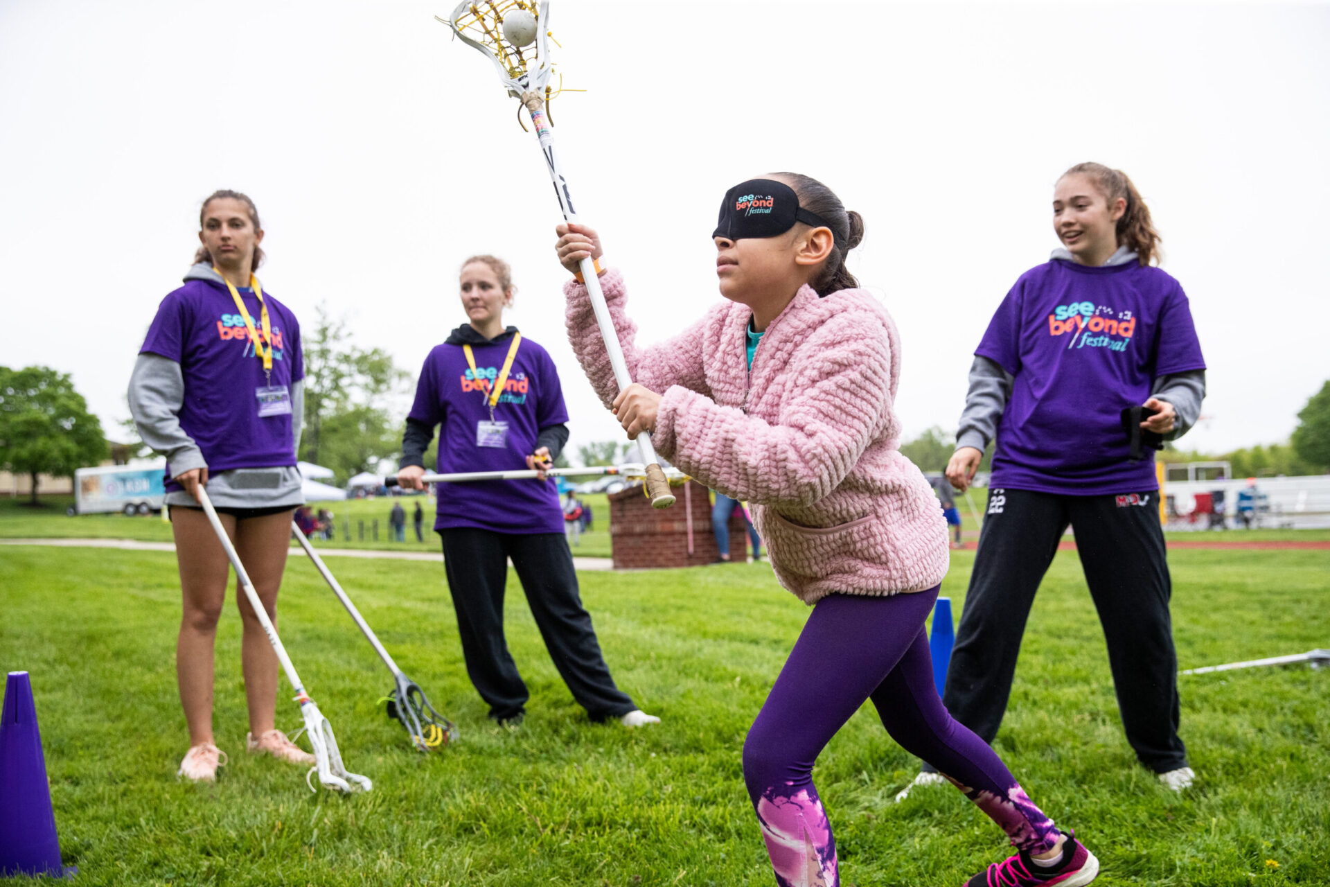 a group of young girls instructing a girl wearing eyeshades on throwing a lacrosse ball from the stick .