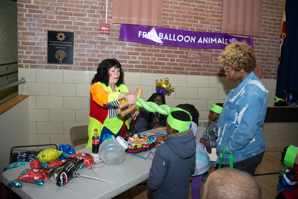 a group of people stand around a table while a clown makes a balloon sword.