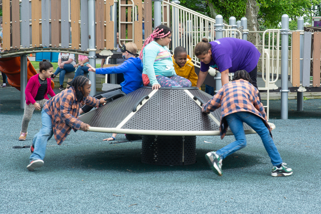 Children play on a metal spinning playground equipment, being pushed by a few kids and attended by an adult in a purple shirt. The playground surface is rubberized, and there's a wooden structure nearby.