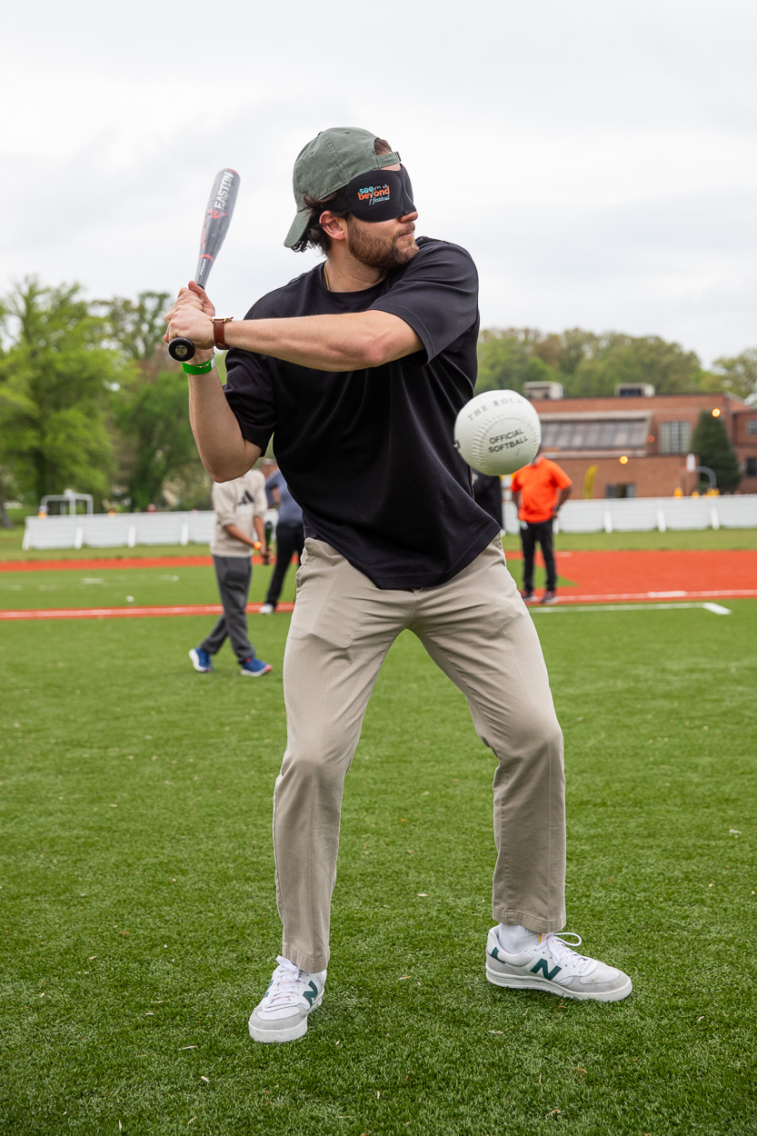 A man wearing eyeshades and a baseball cap prepares to swing a bat at a ball on a grassy field during the See Beyond Festival, with other people and buildings visible in the background.